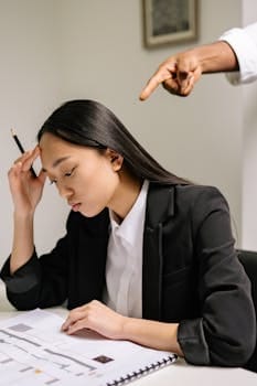 Woman in black blazer studying documents as hand points accusingly, illustrating workplace stress.