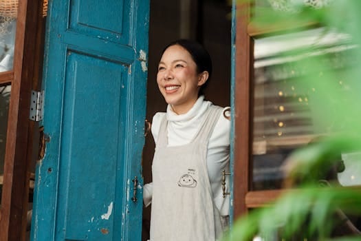 The True Essence of a Home: Why Love Matters More Than Size 3 Joyful woman in apron standing at a rustic blue doorway, exuding warmth and hospitality.