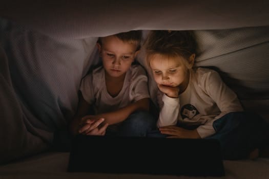 Modern Parenting Challenges And Solutions 3 Two young children watching a tablet under a blanket, creating a cozy nighttime atmosphere.