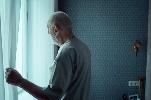 Cutting Off Parents | Why Adult Children Are Choosing Distance Instead of Repair 4 A senior man stands alone, looking out a window with an expression of deep contemplation.